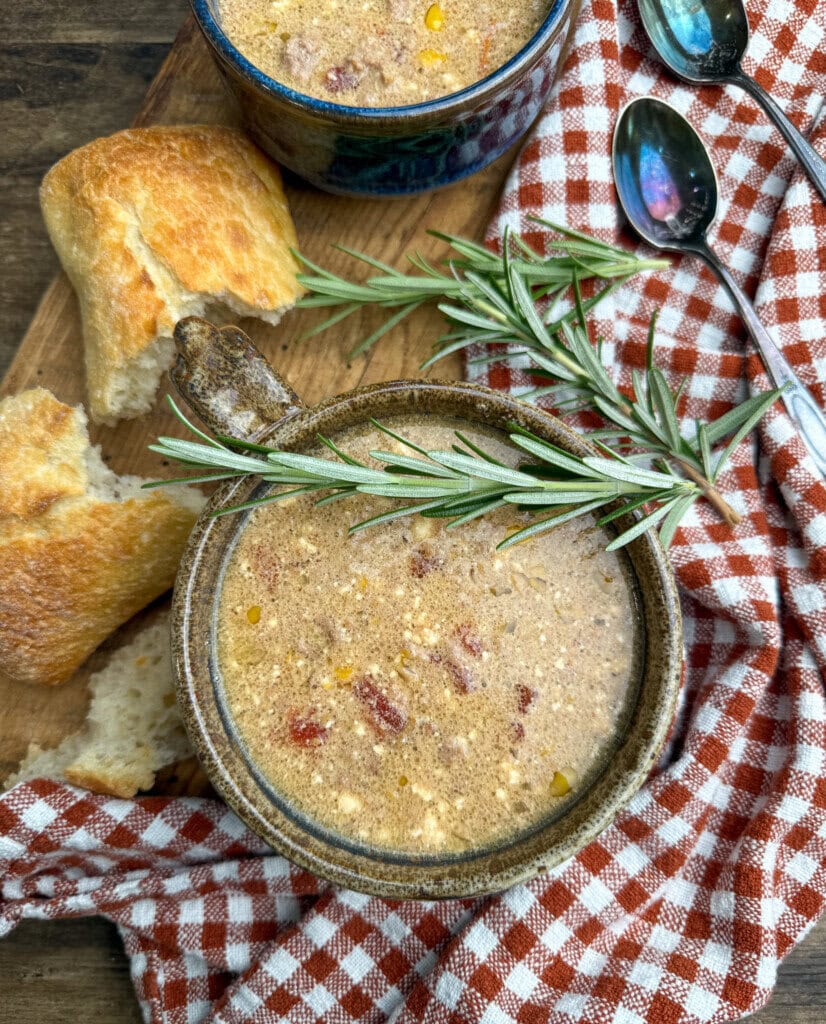 A bowl of creamy soup with herbs and bread on a wooden board, next to a red checkered cloth and two spoons. A sprig of rosemary rests on the bowl.