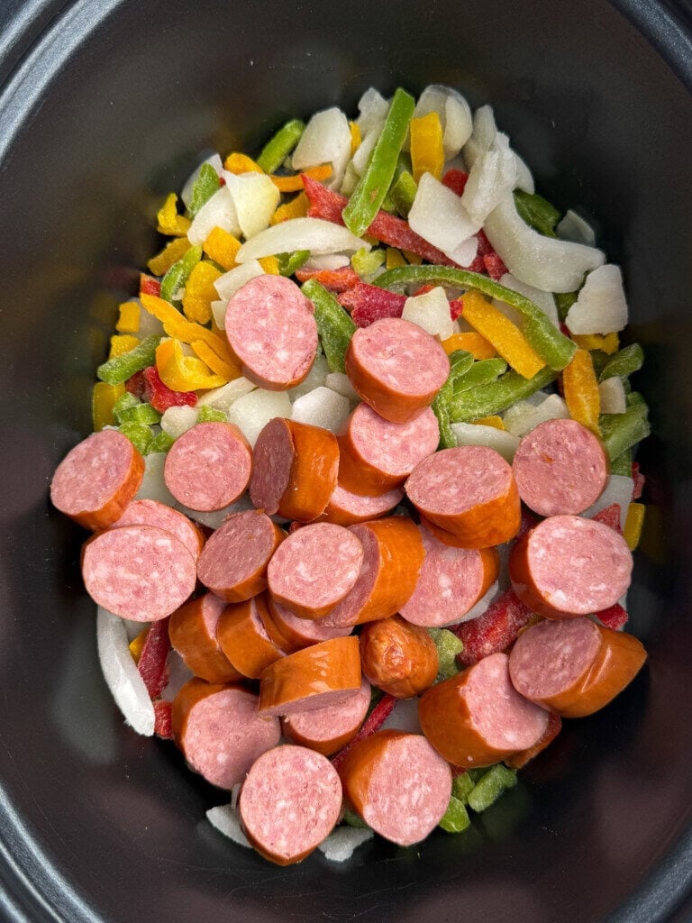 Sliced sausage pieces placed on top of a mixture of chopped green, yellow, and red bell peppers with onions inside a black slow cooker.