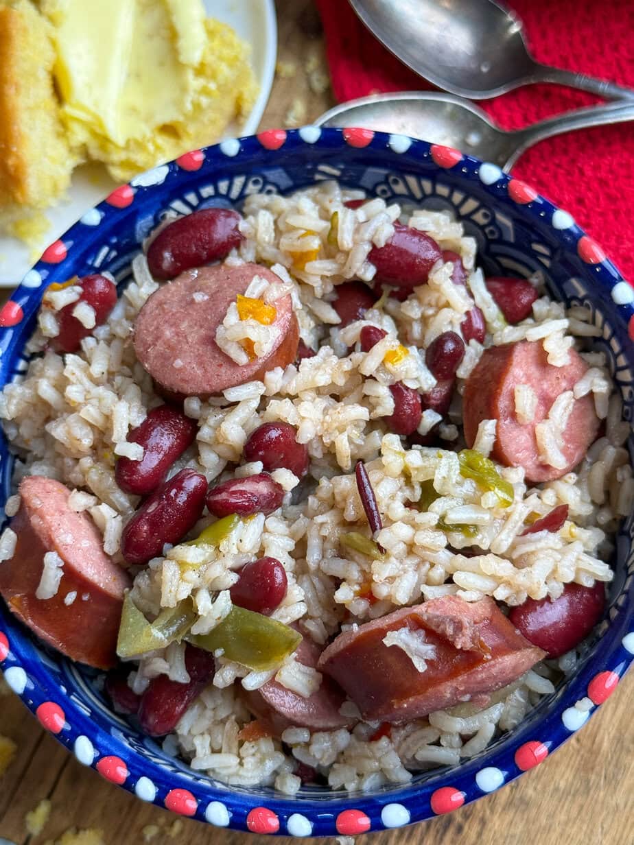 A colorful bowl filled with rice, kidney beans, sliced sausage, and chopped green peppers sits on a wooden surface, with a plate of cornbread and two silver spoons nearby.