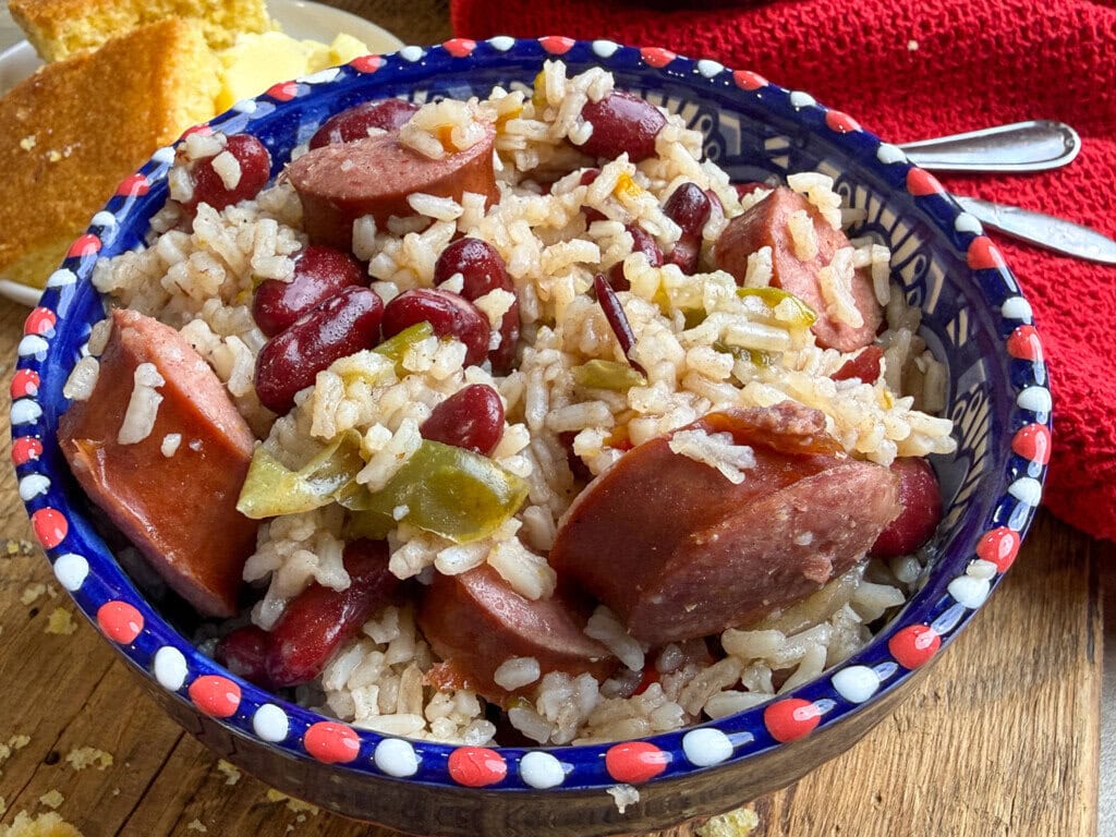 A bowl filled with rice, red beans, sliced sausage, and green peppers sits on a wooden table, with cornbread and a red napkin in the background.