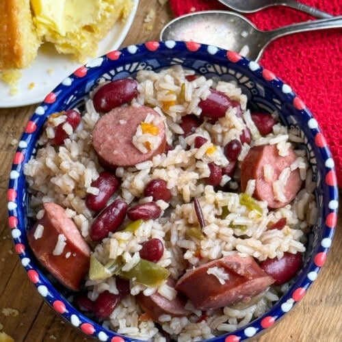 A colorful bowl filled with rice, red beans, sliced sausage, and green peppers on a wooden table next to a plate with buttered cornbread, two spoons, and a red cloth.