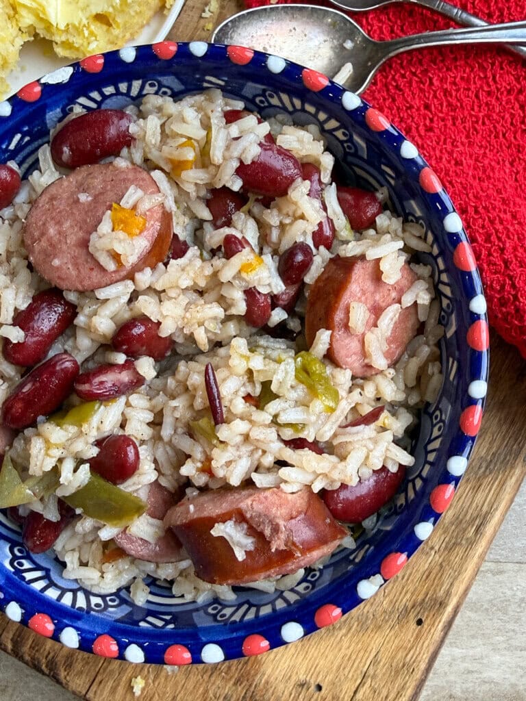 A bowl of rice mixed with sliced sausage, red kidney beans, and chopped green bell peppers, sitting on a wooden board next to a red cloth and two spoons.