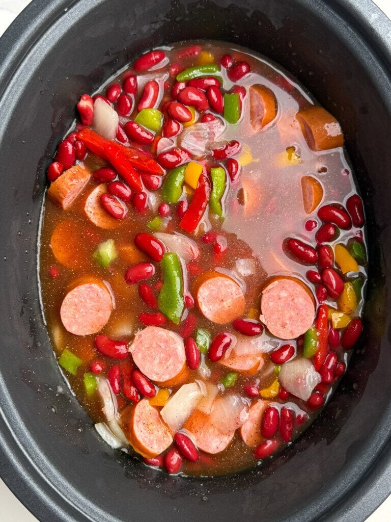 A slow cooker filled with sliced sausage, kidney beans, chopped onions, green and red bell peppers, and broth, ready to be cooked.
