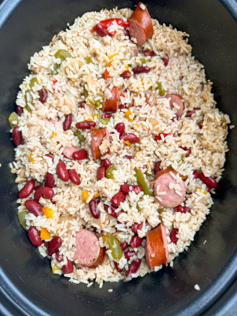 A close-up of cooked rice mixed with red beans, sliced sausage, and diced green and yellow peppers in a black pot.
