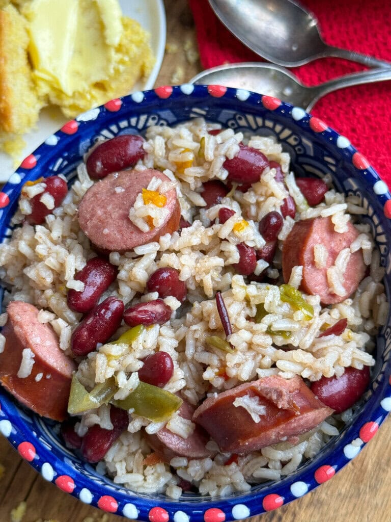 A colorful bowl filled with rice, kidney beans, sliced sausage, and green bell peppers. In the background, a plate with buttered cornbread and two metal spoons on a red cloth are partially visible.
