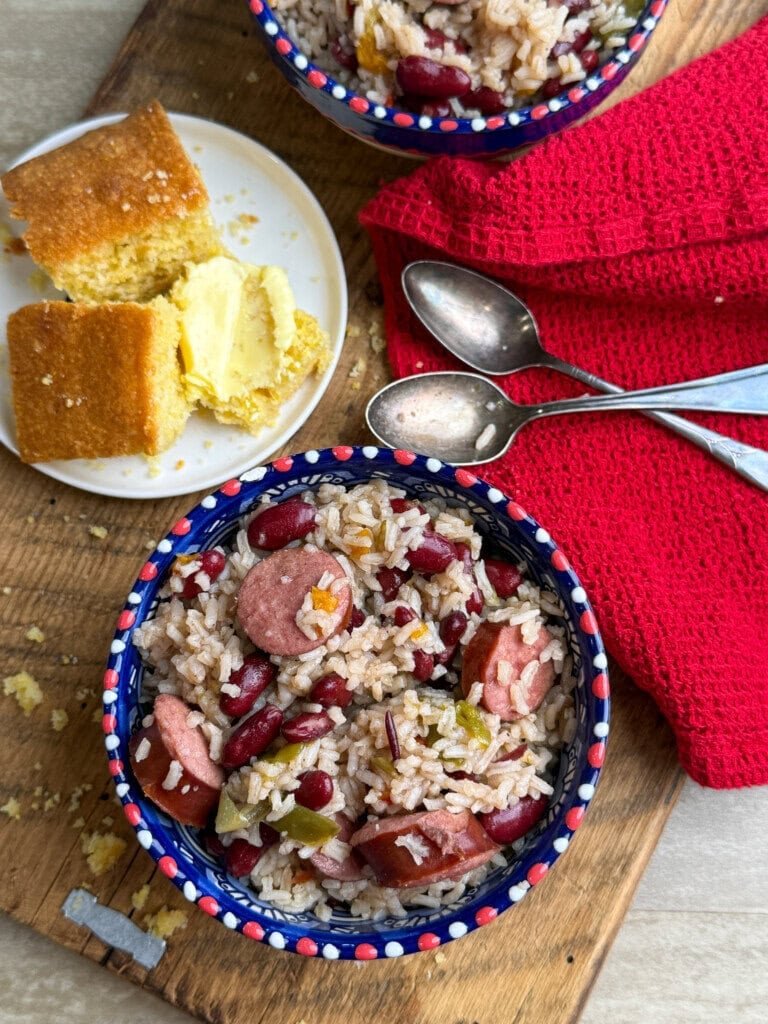 A bowl of rice with sausage, kidney beans, and vegetables sits on a wooden board next to buttered cornbread, three spoons, and a red cloth napkin. Another bowl of the rice dish is partially visible at the top.