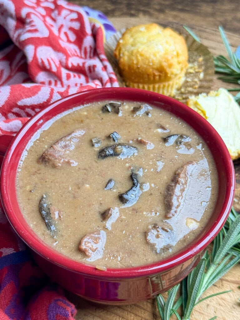 A red bowl filled with brown bean soup containing beef stew and mushrooms sits on a wooden table, next to a cornbread muffin, rosemary, and a red patterned cloth.