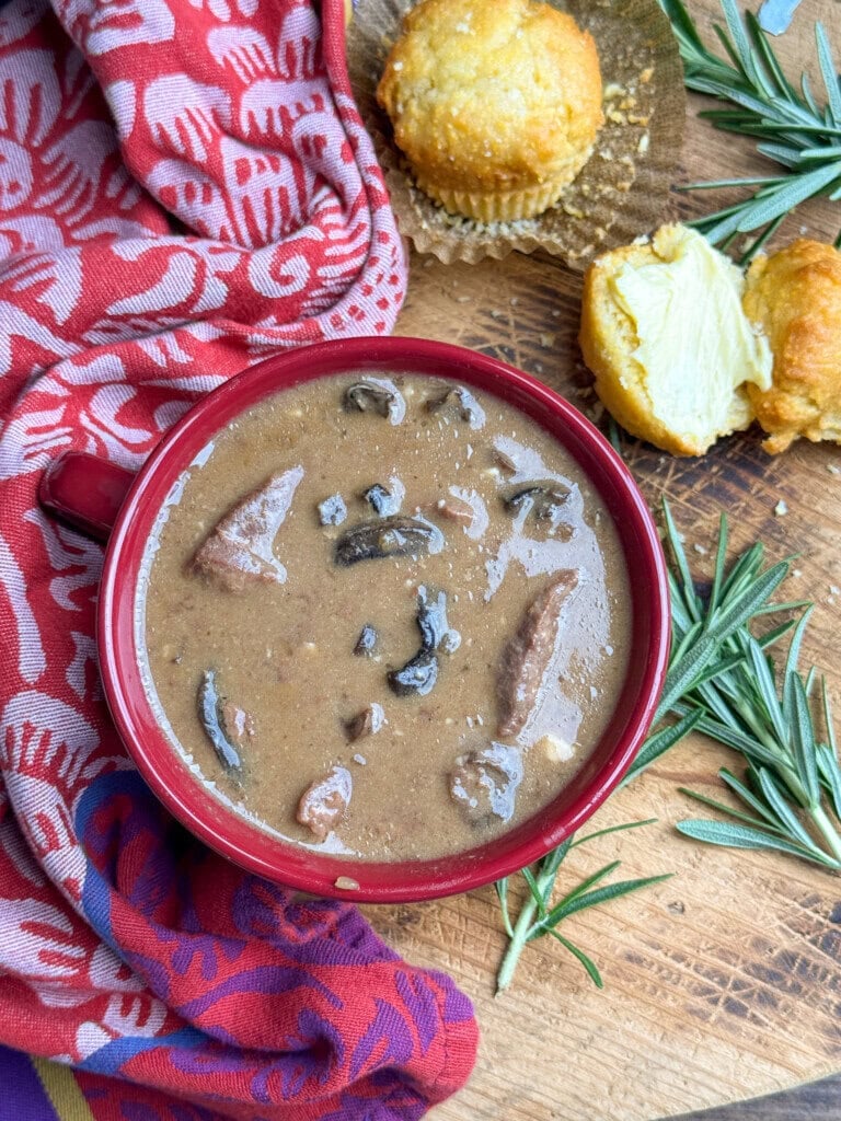 A red bowl of beef and mushroom stew sits on a wooden board with two cornbread muffins, fresh rosemary, and a red patterned cloth beside it.