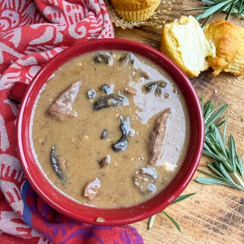 A bowl of brown mushroom and meat stew sits on a wooden surface beside split cornbread muffins, rosemary sprigs, and a red patterned cloth.