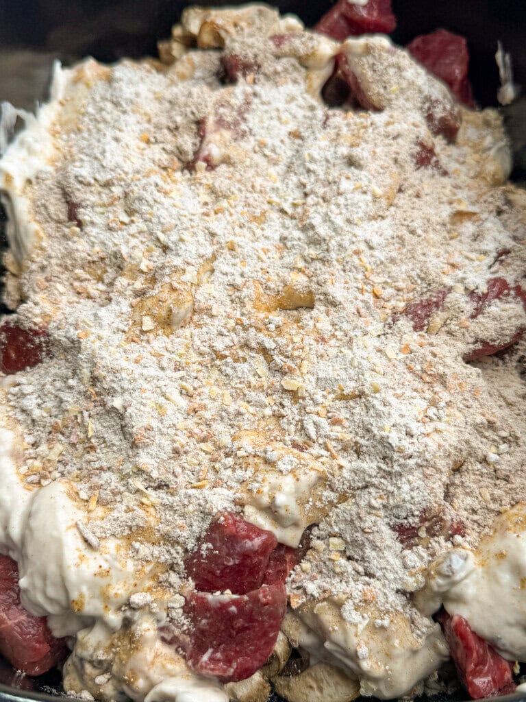 Close-up of an uncooked beef stew with chunks of raw meat, creamy sauce, and a layer of seasoning on top