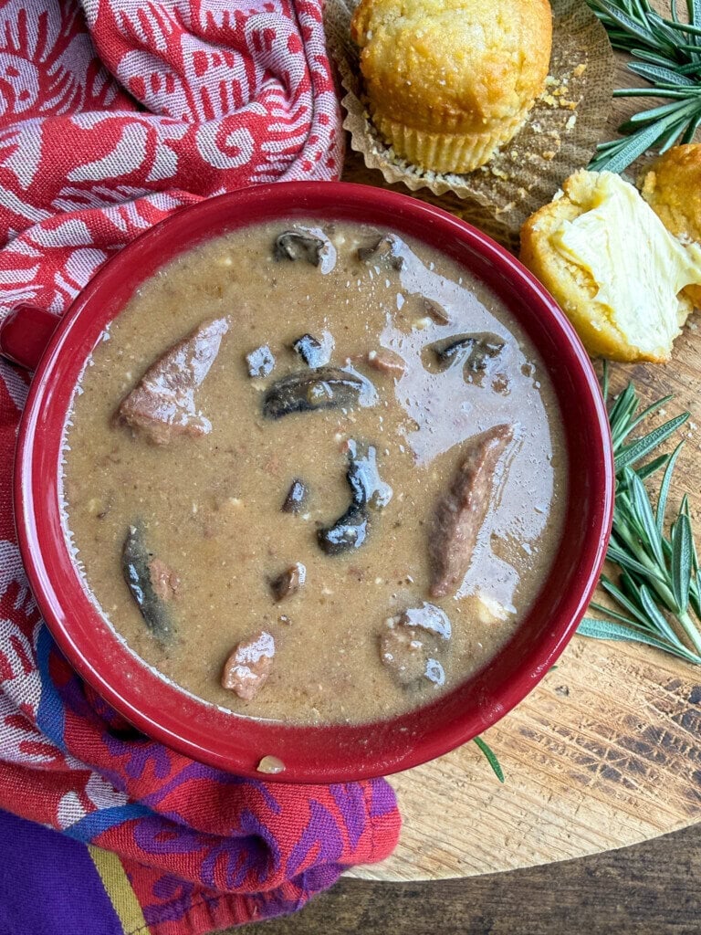 A bowl of brown stew with beef chunks and mushrooms, placed on a wooden surface next to a muffin, a rosemary sprig, and a red patterned cloth.
