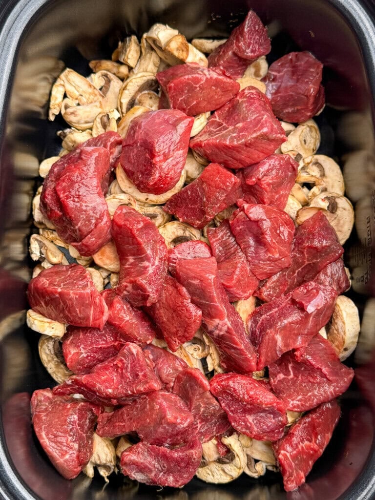 Chunks of raw beef are layered over sliced mushrooms in a black slow cooker insert, ready to be cooked.