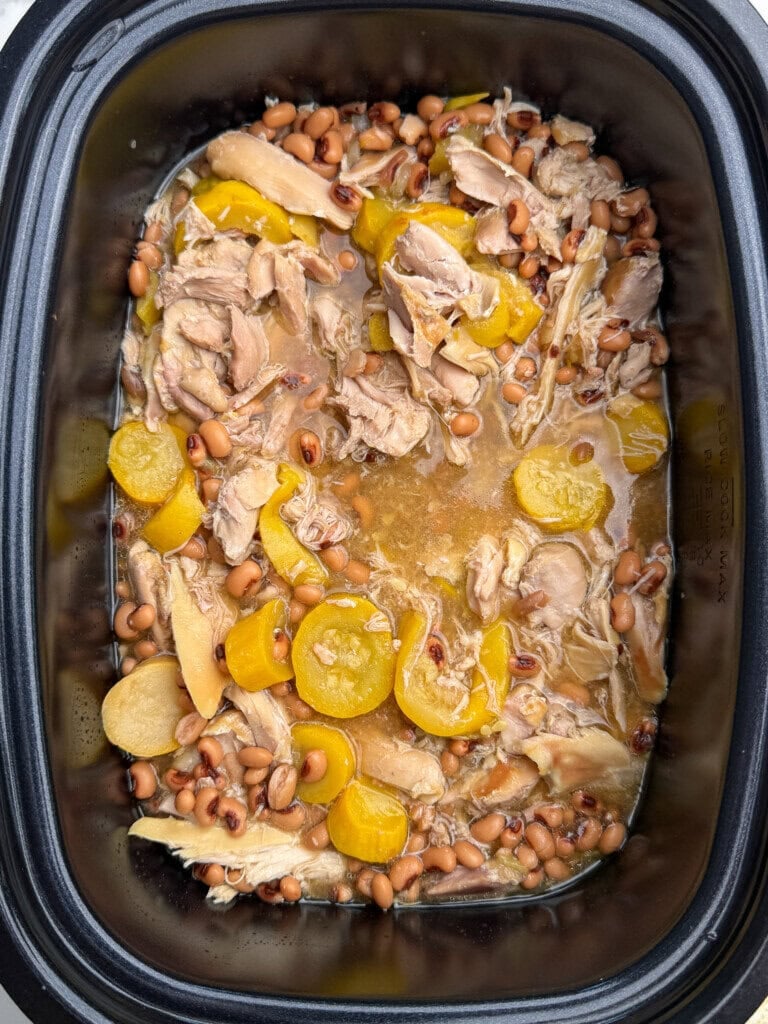 Cooked chicken, black-eyed peas, and sliced yellow squash in a slow cooker with broth. The ingredients appear tender and well-mixed, filling most of the cooking pot with visible liquid at the bottom.