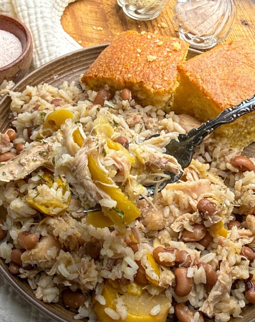 A plate of chicken and rice with black-eyed peas and yellow squash, served with cornbread on the side. A fork rests on the plate, and salt and pepper shakers are in the background.