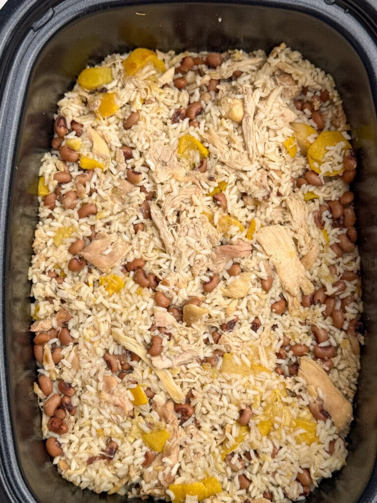 A close-up of a dish in a black slow cooker, featuring rice, shredded chicken, black eyed peas, and pieces of yellow squash