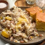 A plate with rice, shredded chicken, black-eyed peas, squash, and two pieces of cornbread. Salt and pepper shakers and a bowl of salt are in the background. The plate is on a wooden table with a white cloth nearby.