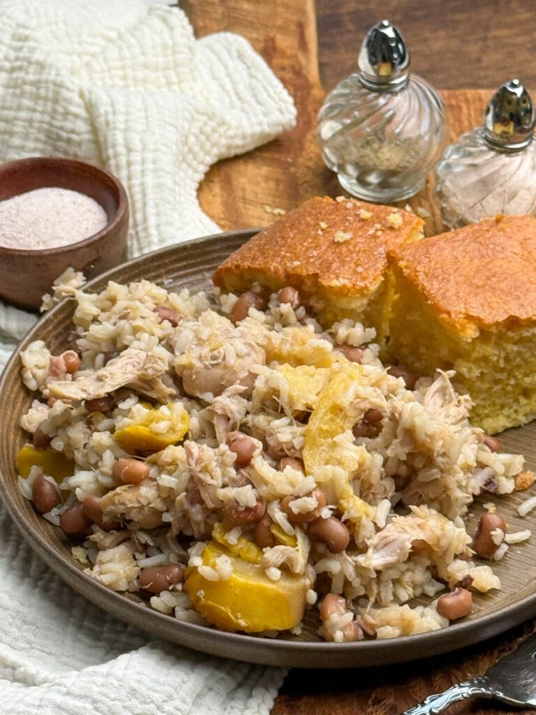 A plate with rice, black eyed peas, shredded chicken, yellow squash, and two pieces of cornbread. Salt and pepper shakers and a small bowl of salt are in the background on a wooden surface.