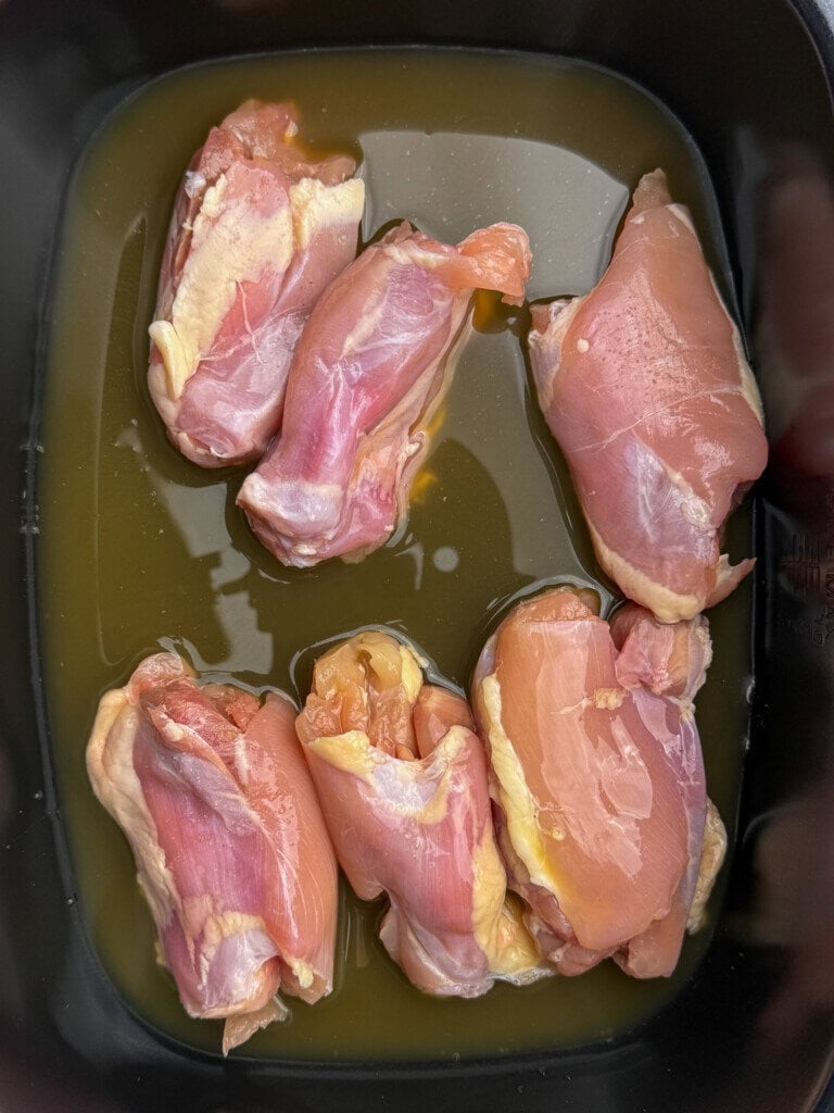 Six pieces of raw chicken sitting in a shallow pool of yellowish marinade or liquid inside a black crock pot