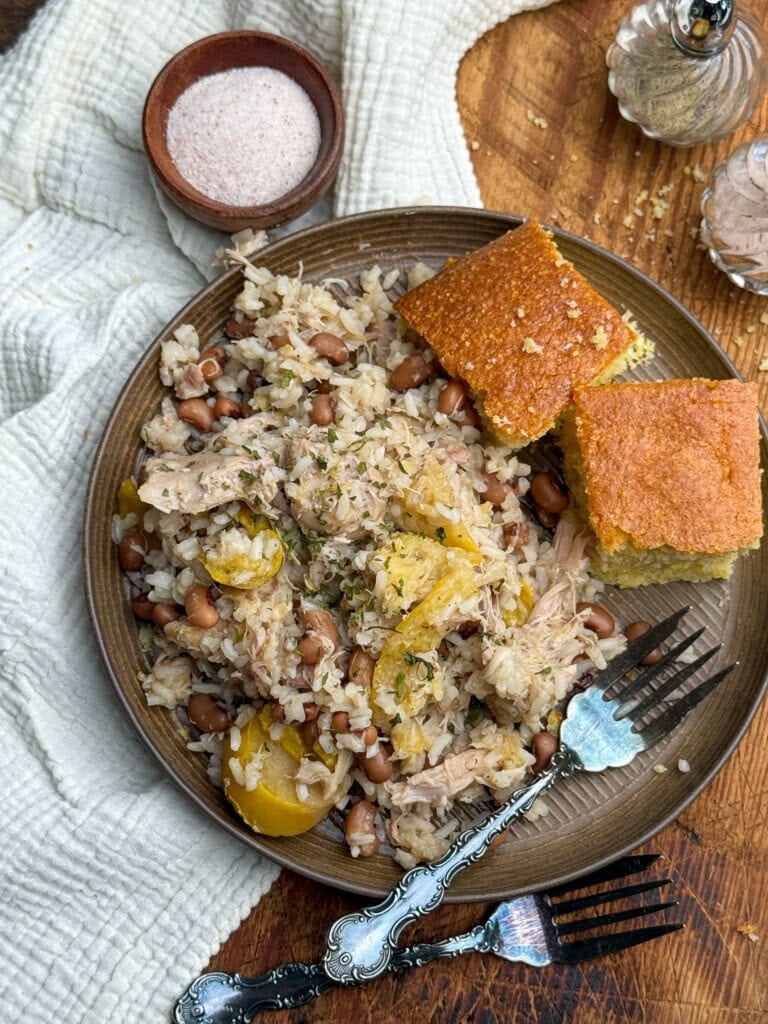 A plate of rice with beans, shredded meat, and yellow squash, served with two pieces of cornbread. A bowl of salt and a pepper shaker sit nearby on a wooden table with a white cloth.