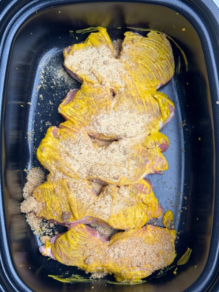 Raw pork rib pieces coated with yellow mustard and sprinkled with brown sugar are arranged in a black roasting pan, ready to be cooked.