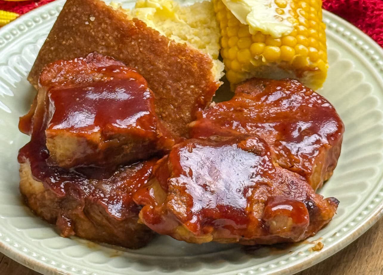 A plate with two pieces of barbecue-glazed meat, a square of cornbread, and a piece of corn on the cob topped with melting butter.