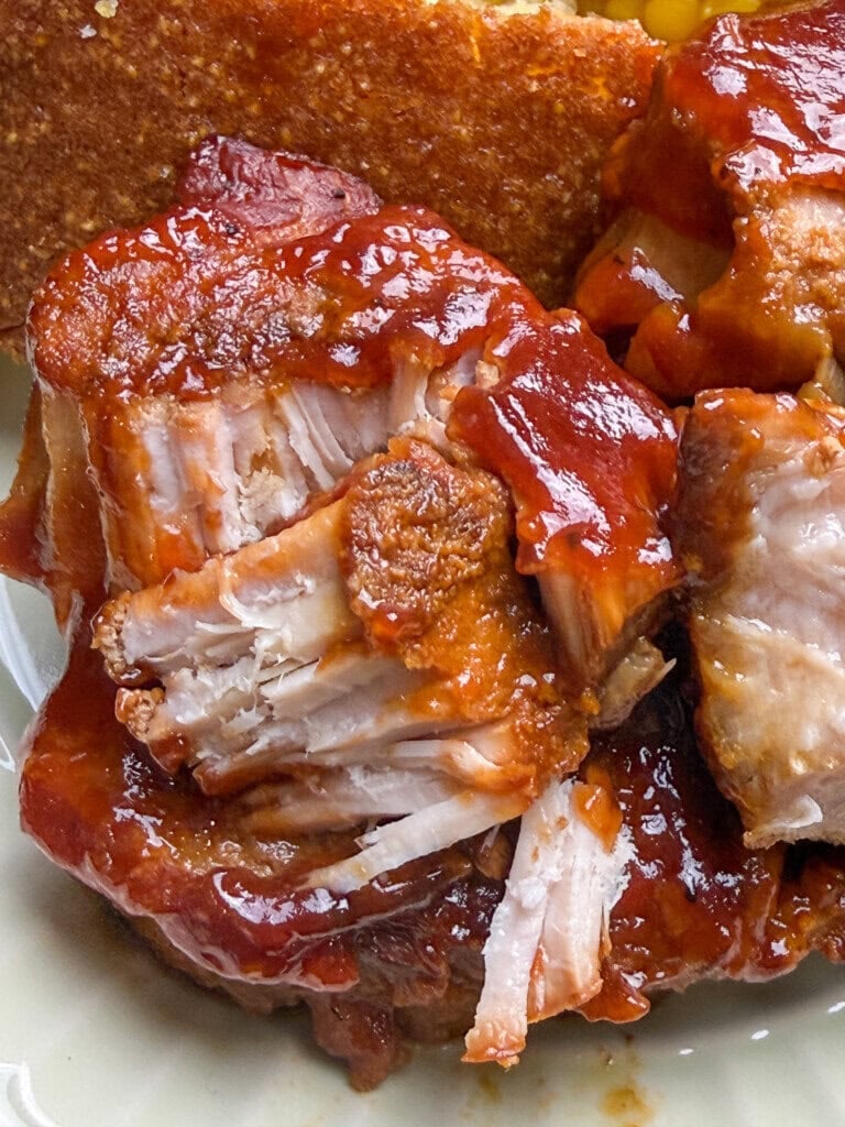 Close-up of tender, saucy barbecue ribs with a side of cornbread in the background. The ribs are coated in a thick, glossy barbecue sauce and look juicy and flavorful.