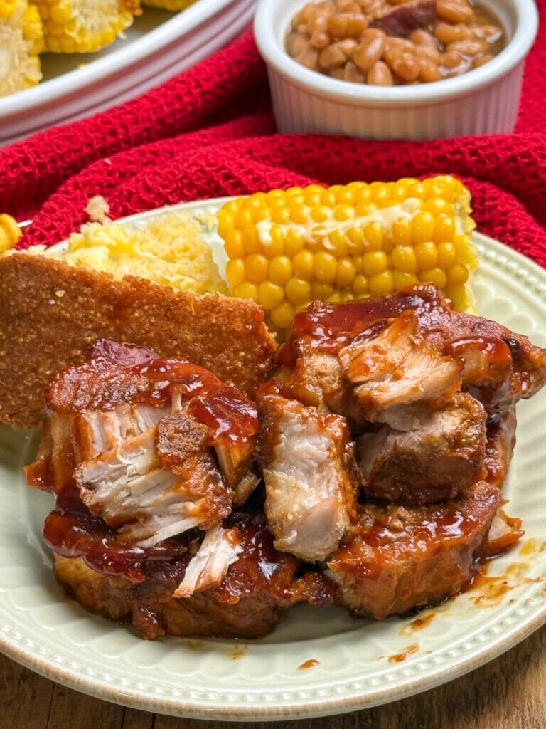 A plate with barbecue ribs, cornbread, and corn on the cob. In the background, baked beans in a white bowl and a red cloth are visible.