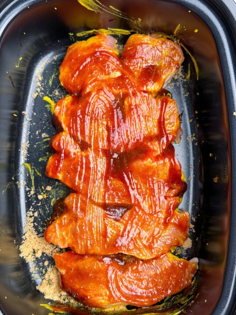 Raw pork ribs coated in a reddish-orange BBQ sauce and sprinkled with brown sugar, arranged in a black roasting pan, ready for cooking.