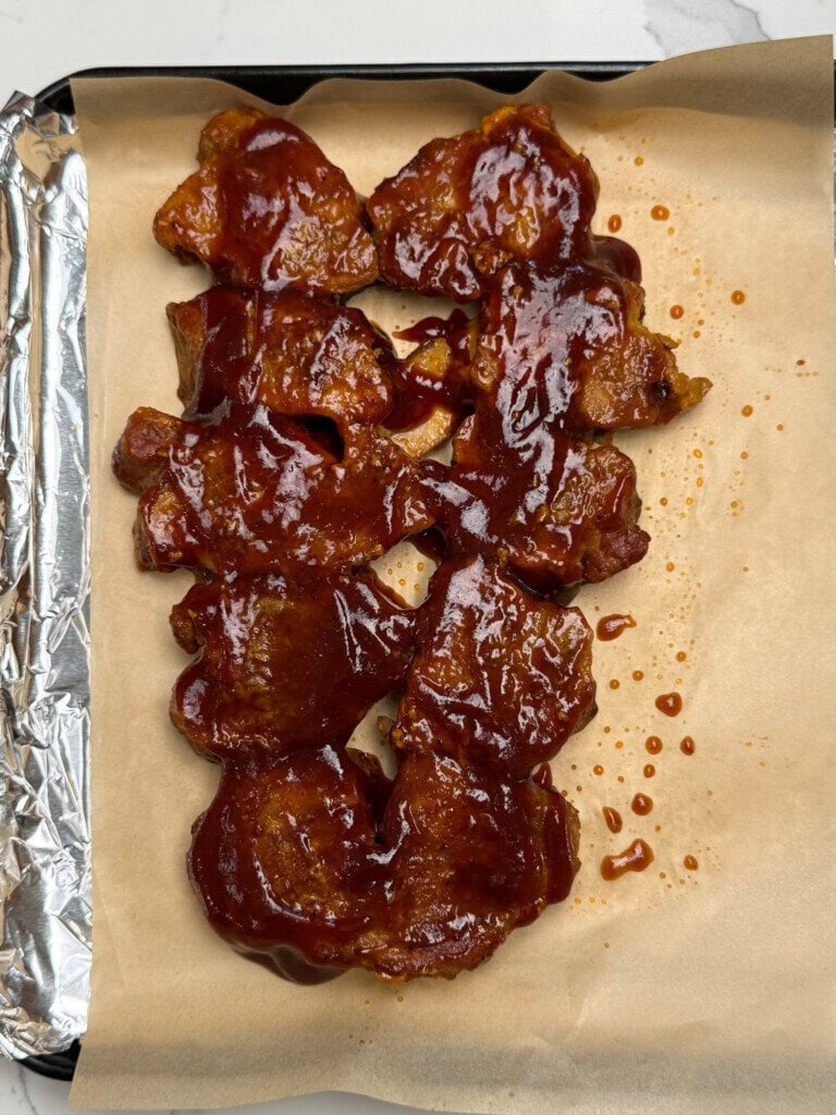 Barbecue sauce-covered pieces of meat are arranged on parchment paper atop a baking tray, with part of the tray wrapped in aluminum foil.