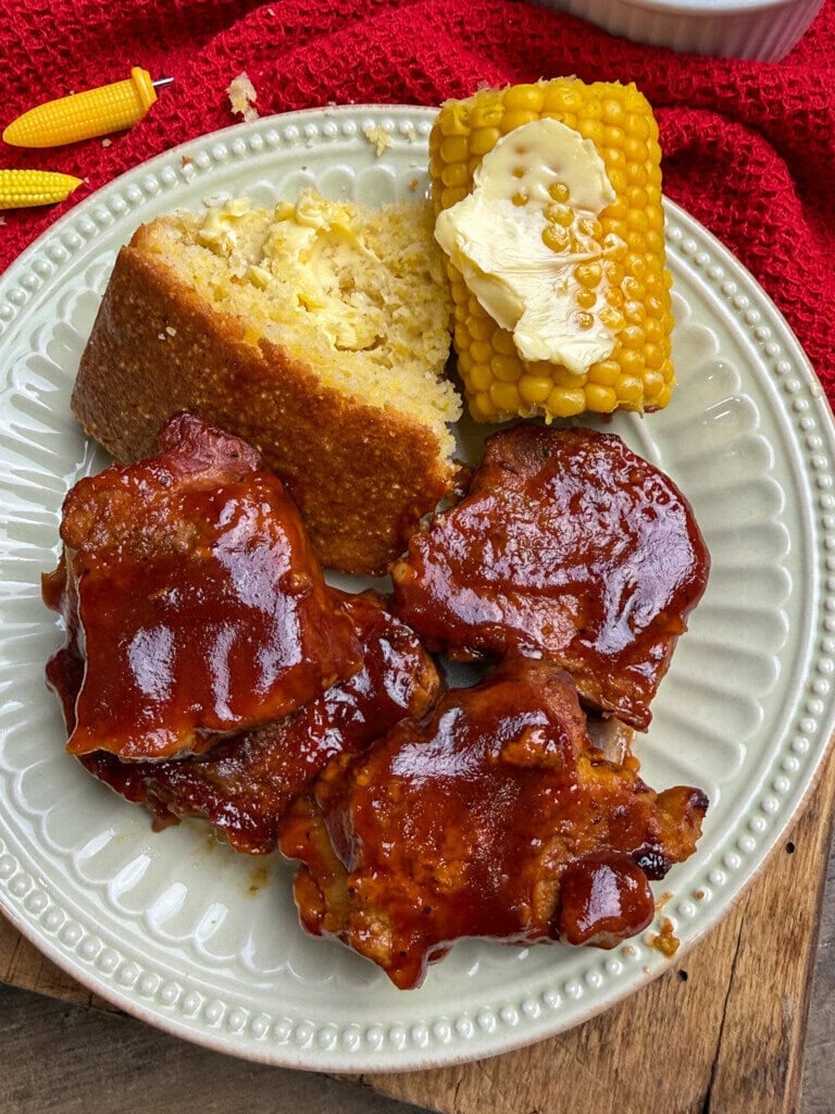 A plate with three pieces of barbecue ribs, a slice of cornbread, and corn on the cob topped with butter, set on a white plate with a red cloth in the background.