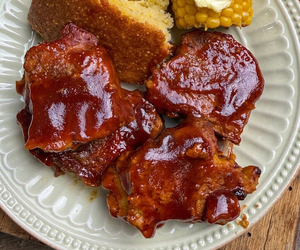 Three pieces of barbecue-glazed country-style pork ribs sit on a plate with a slice of cornbread and buttered corn on the cob.