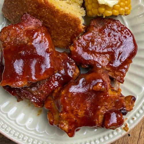 Three pieces of barbecue-glazed country-style pork ribs sit on a plate with a slice of cornbread and buttered corn on the cob.