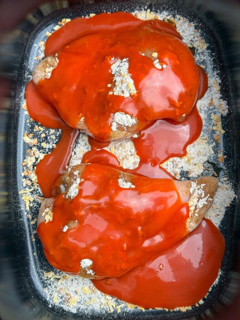 Two pieces of chicken covered in bright red dressing, placed in a black crock pot with some dry onion soup mix scattered around.