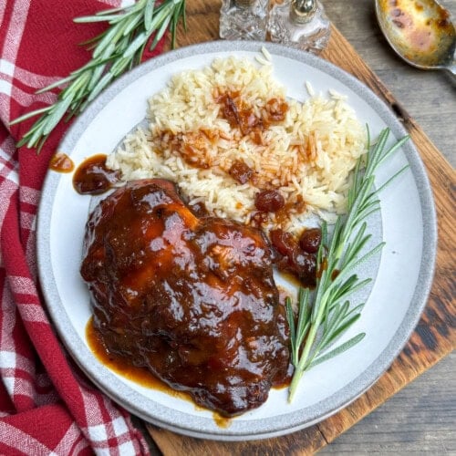 A plate with saucy cranberry chicken, white rice topped with sauce, and fresh rosemary sprigs, set on a wooden board next to a red checkered cloth and salt and pepper shakers.