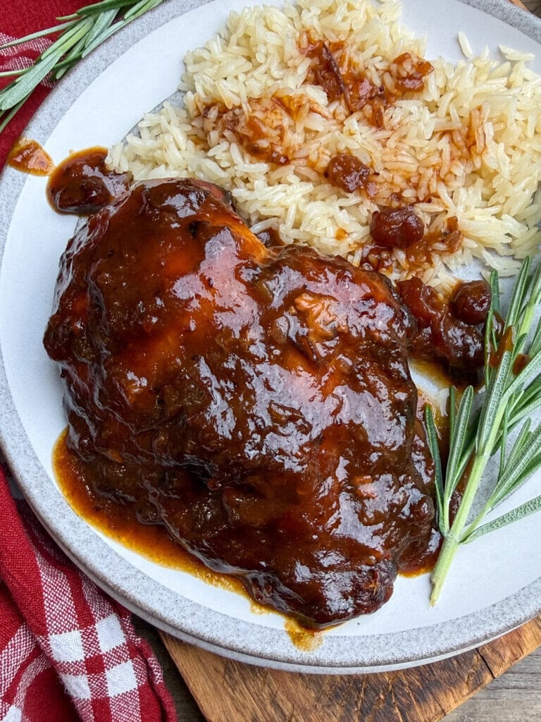 A plate with glazed cranberry chicken, white rice topped with sauce, and sprigs of fresh rosemary, set on a wooden surface with a red checkered napkin nearby.