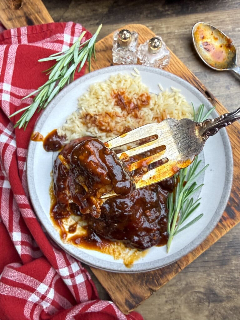 A fork holds a bite of saucy chicken over a plate with rice and meat covered in rich brown sauce, garnished with rosemary. A red checkered cloth and a spoon with sauce are beside the plate on a wooden surface.