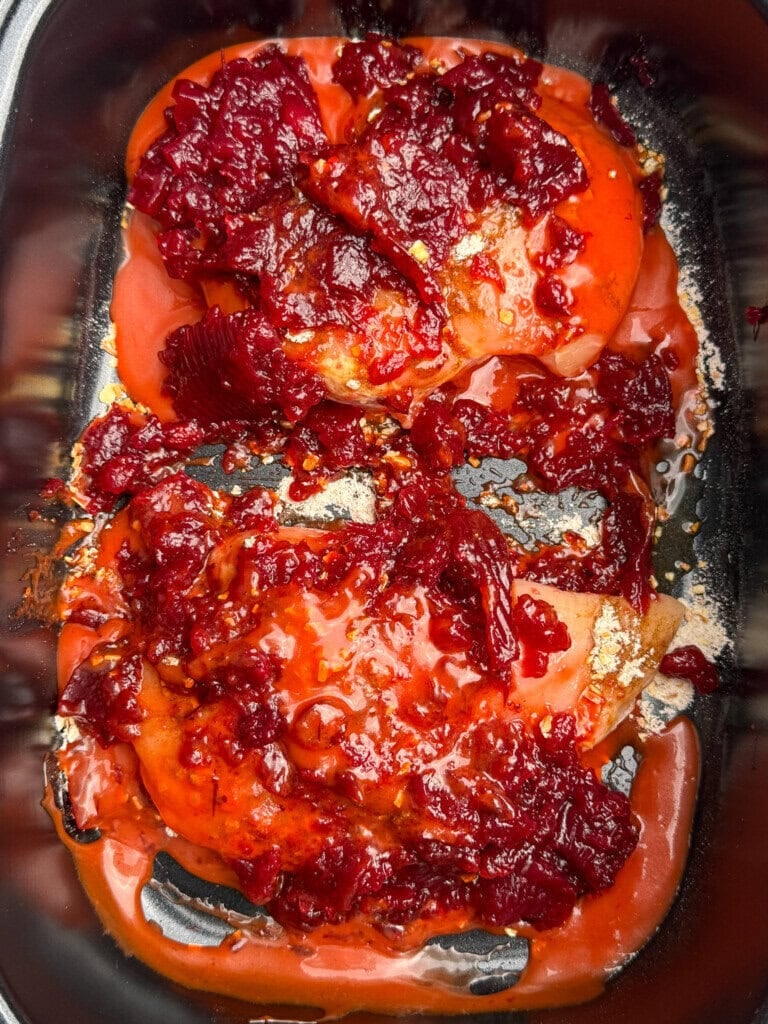 Two raw chicken breasts in a black crock pot, topped with cranberry sauce.