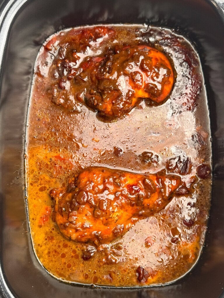 Two pieces of chicken in a dark, rich sauce with visible spices and bits of vegetables, served in a black rectangular dish. The sauce has an oily sheen and reddish-brown color.