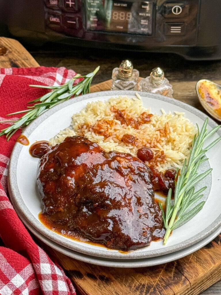 A plate with glazed chicken in rich sauce, white rice topped with sauce, and garnished with rosemary sprigs. A slow cooker, salt and pepper shakers, and a red checkered napkin are in the background.
