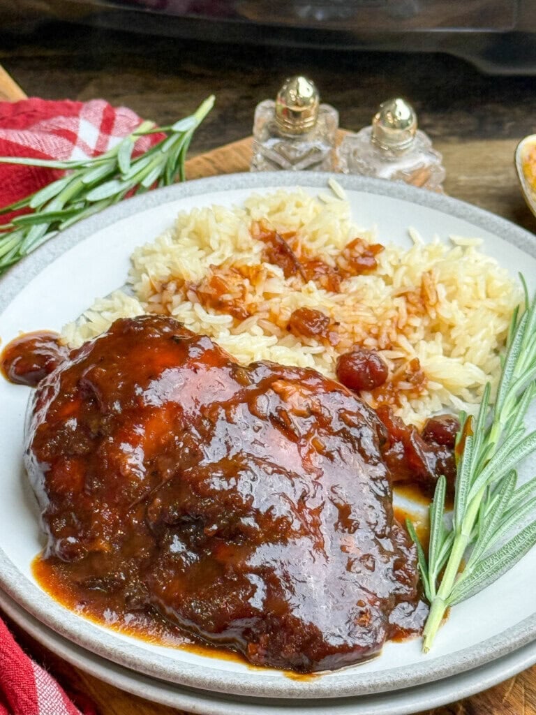 A plate of glazed, saucy chicken served with rice, garnished with fresh rosemary, with salt and pepper shakers and a red cloth napkin in the background.
