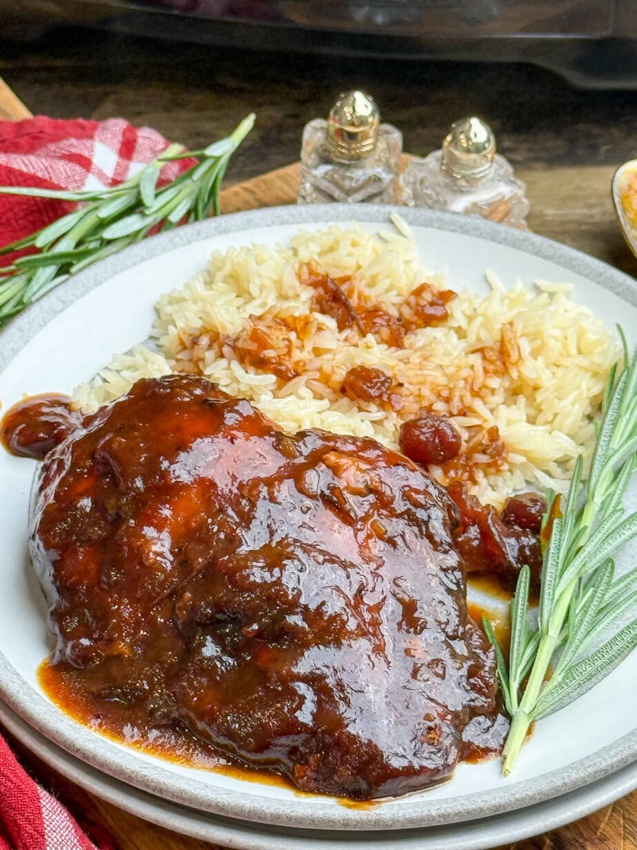 A plate of glazed, saucy chicken served with rice, garnished with fresh rosemary, with salt and pepper shakers and a red cloth napkin in the background.