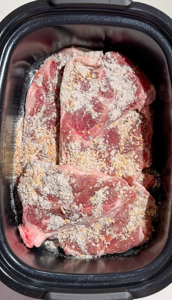 Two raw pork steaks in a slow cooker, topped with a coating of dry seasoning and onion soup mix. The meat appears marbled, and the spices are spread evenly across the surface.
