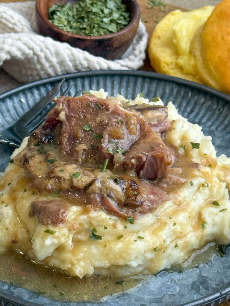A plate of mashed potatoes topped with shredded pork steaks and gravy, garnished with herbs. A fork rests on the plate, with bread and a bowl of dried herbs in the background.