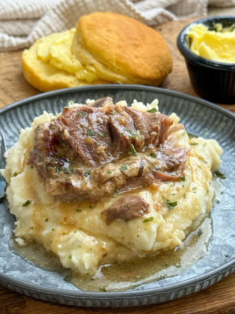 Tender pork steaks with gravy over mashed potatoes on a plate, garnished with herbs. In the background, there is a biscuit with butter and a small dish of butter.