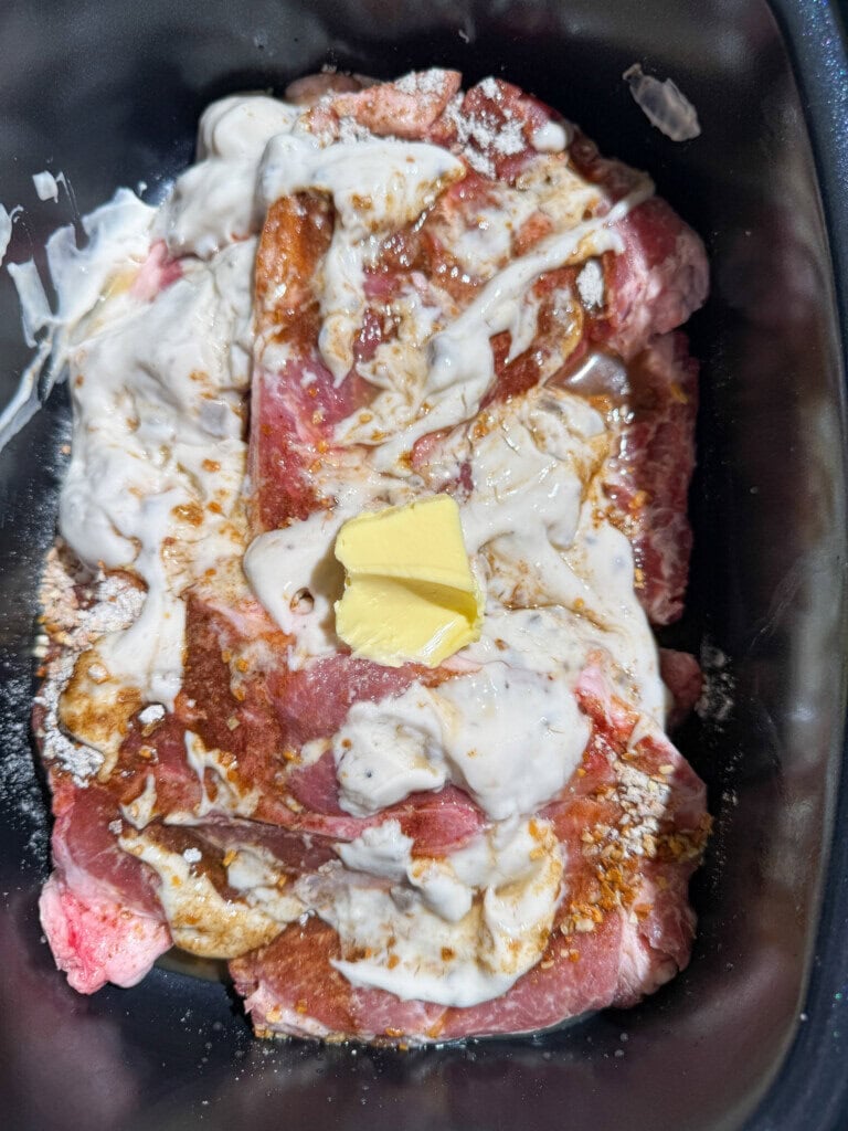 Raw pork covered with condensed mushroom soup, seasonings, and a pat of butter sits in a black slow cooker, ready to be cooked.