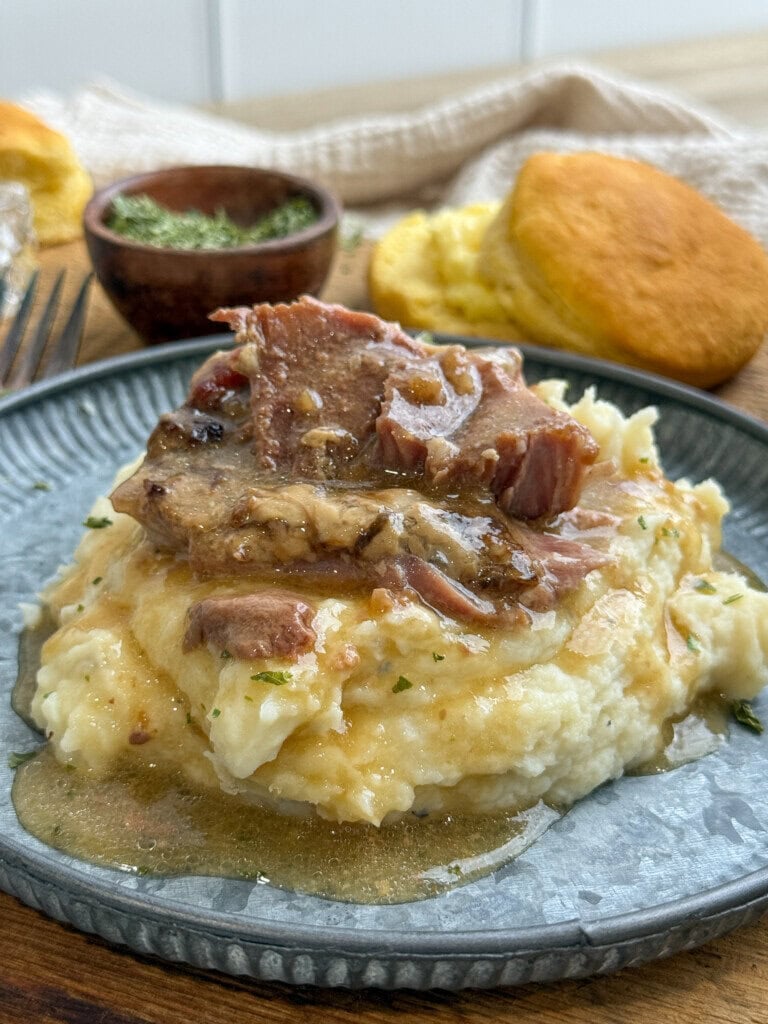 A plate of mashed potatoes topped with tender meat and gravy, with a biscuit and a small bowl of herbs in the background on a rustic wooden table.