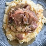 A serving of tender meat with gravy sits on a bed of mashed potatoes, garnished with chopped herbs, on a gray plate.