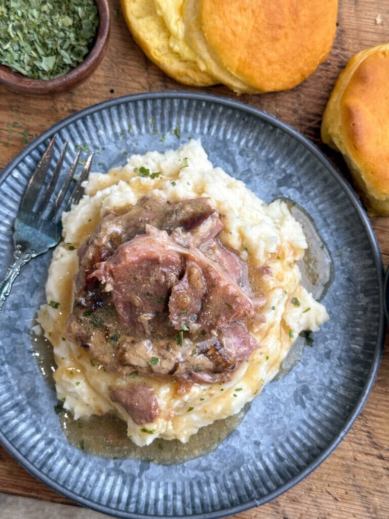 A plate of mashed potatoes topped with pork steaks and gravy, garnished with herbs. A fork rests on the plate, and golden biscuits are on the wooden surface nearby.