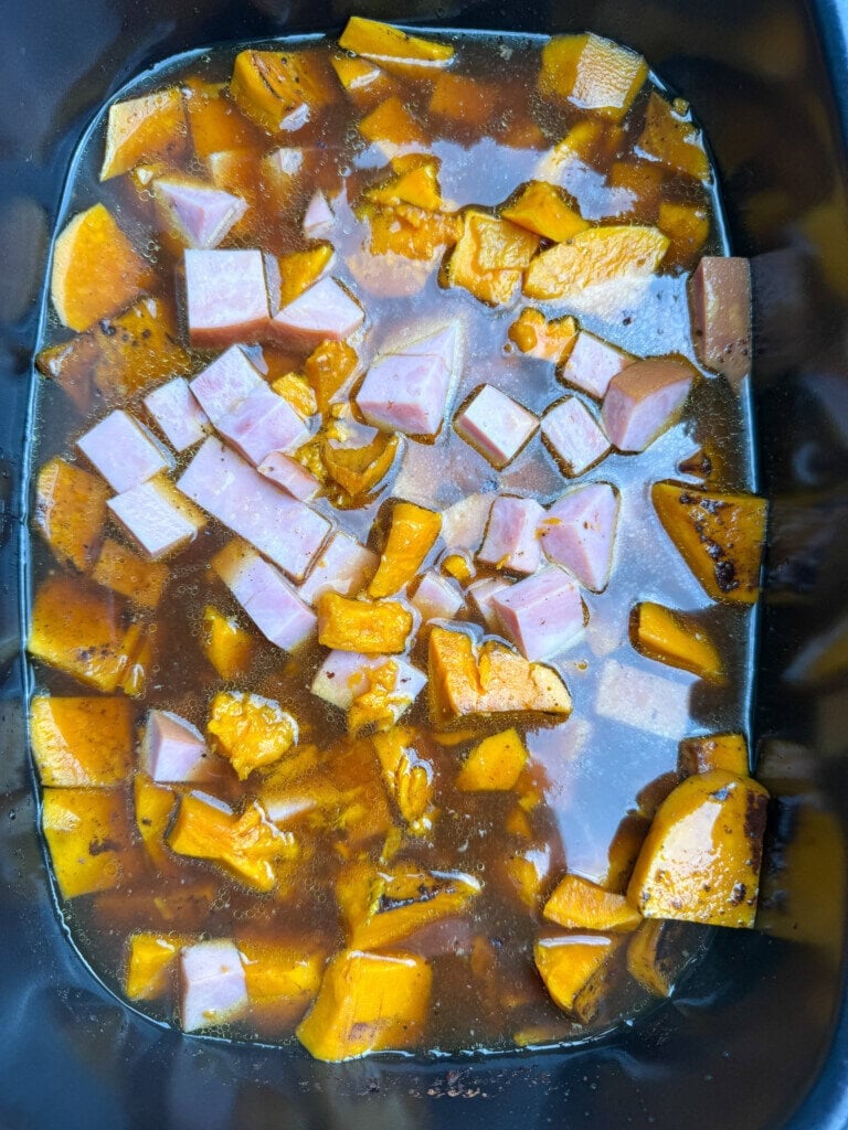 Cubed ham and chunks of sweet potatoes in broth inside a black slow cooker, ready for cooking.