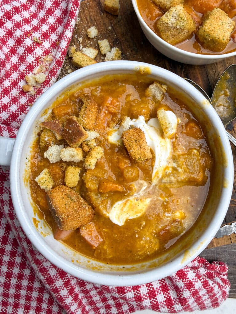 A bowl of thick sweet potato soup topped with croutons and a swirl of cream sits on a wooden surface next to a red and white checkered cloth. Crumbs and a spoon are nearby, with another bowl of soup in the background.